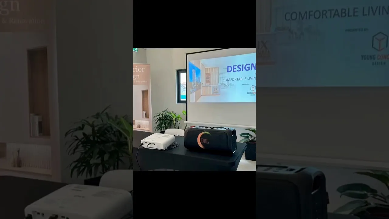 A projector screen reading “Design, Comfortable Living” set up in the IX SURIA sales gallery for the Home Talk Series, with a speaker and potted plants on the table